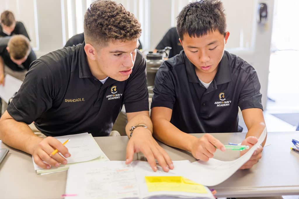 Two students in black polo shirts sit at a table, working together on paperwork.