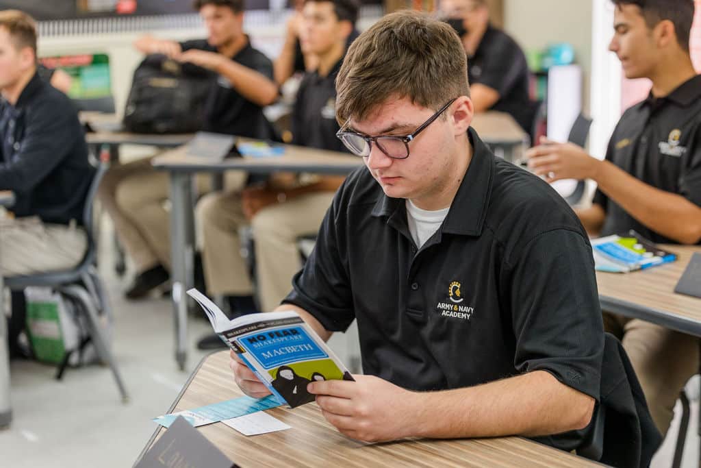 A student in a black polo shirt and glasses reads a book at a classroom desk.