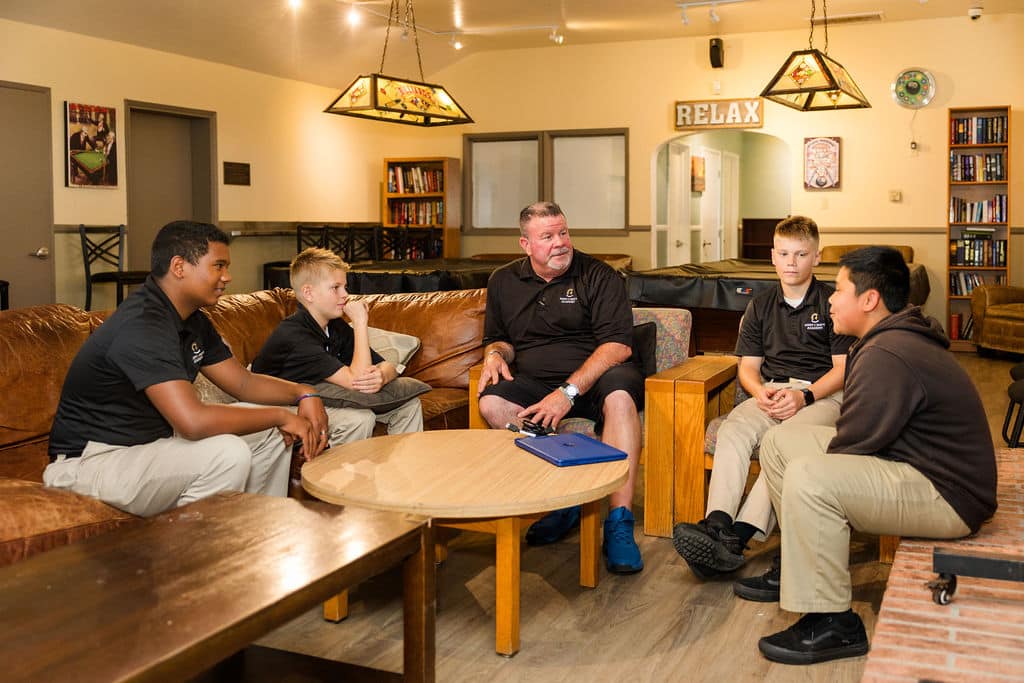 Five people engaged in a communication sit on couches around a wooden table in a relaxed lounge setting.