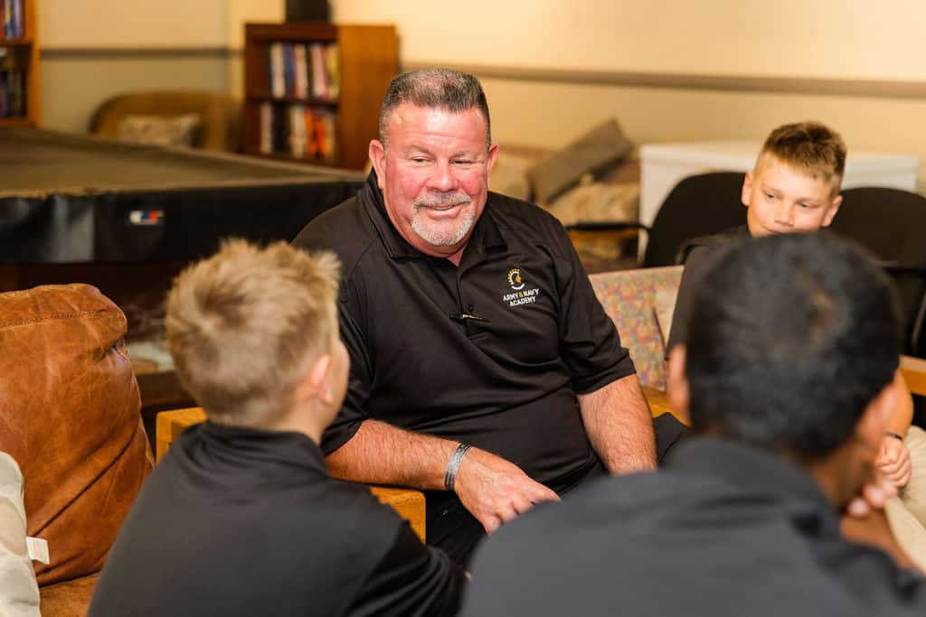 A mentor in a black shirt is sitting and talking with a group of children