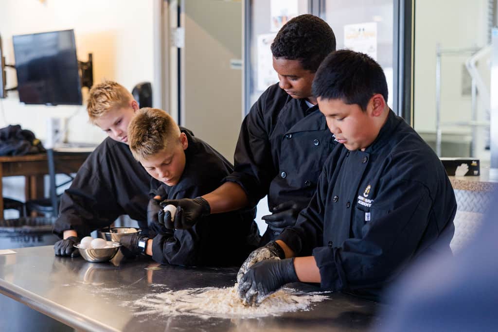 Four young people in black chef uniforms work together in a kitchen.