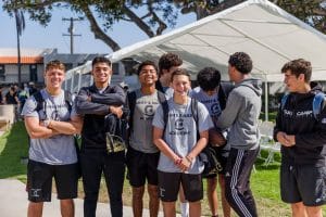 A group of teenage boys in 'Army & Navy Academy' shirts stand outdoors, smiling and enjoying a sunny day.