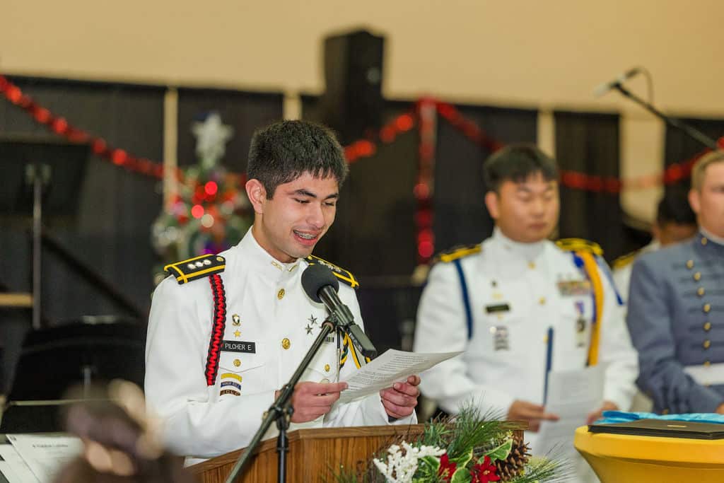 A young man in a formal white military uniform speaks at a podium with a microphone, holding a piece of paper.