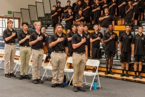 Students standing in a gymnasium, with hands over their hearts, participating in a formal ceremony.